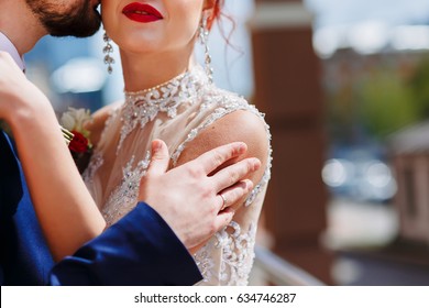 A Young Man Gently Embraces His Bride In A Wedding Dress By The Shoulders To The Goosebumps. Bride & Bridegroom. Groom Kisses Bride's Neck. Girl With Red Lips.