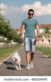 Young Man And German Spitz Walk In The Park - He Keeps The Dog On The Leash