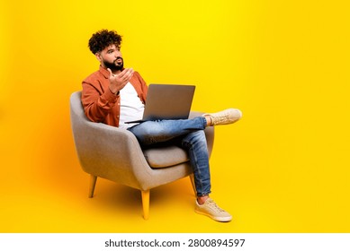 A young man gestures while seated in a gray armchair with a laptop on his lap against a bright yellow backdrop, conveying remote work, creativity, casual conversation and modern studio vibes.