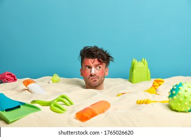 Young Man Gets Suntan At Beach, Buried In Sand, Has Sunburn Face Skin, Applies Sunnscreen On Nose, Poses At Seaside With Various Items For Rest, Looks Aside. People, Vacation, Holiday, Relax Concept