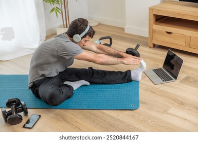 Young man getting fit doing sports with headphones, stretching, yoga, calisthenics in the living room of his minimalist home with online personal trainer, healthy living, training routines at home