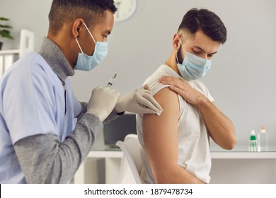 Young Man Getting Flu Or Covid-19 Antiviral Shot During Vaccination Campaign. African-American Doctor In Face Mask Holding Syringe And Cleaning Skin On Patient's Arm Before Antivirus Vaccine Injection