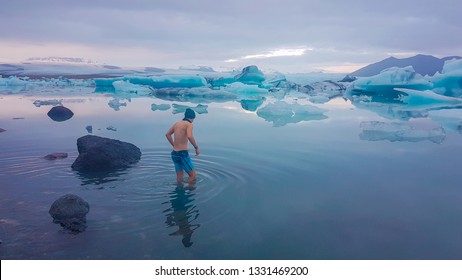 Young man getting into the water of Glacier lagoon. Man wearing only swimming shorts and a hat. Ice bergs drifting in the lagoon. Cold temperatures for ice swimming. Calm surface of the water.