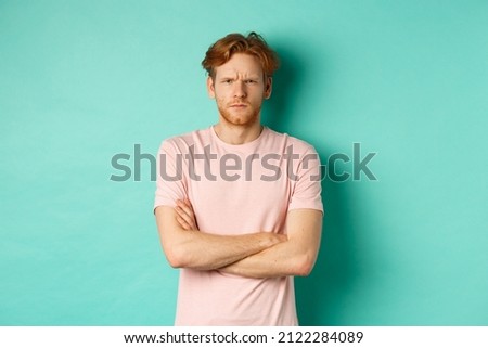 Young man with ginger hair and beard looking offended, feeling insulted and staring at camera, sulking at you, cross arms on chest defensive, standing over turquoise background
