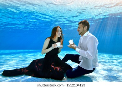 Young Man And Girl Sitting Under Water At The Bottom Of The Pool With White Cups In Their Hands, Smiling And Looking At Each Other. A Girl In A Dress, A Guy In A White Shirt. Tea Party Underwater