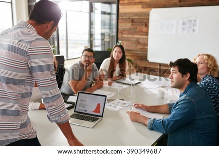 Young man giving business presentation on laptop to colleagues sitting around table in conference room.