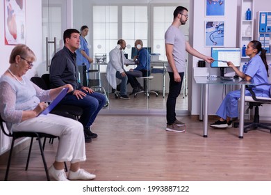 Young Man Giving Radiography To Assitant Making New Appointment Using Computer. Crowded Hospital Waiting Area With People Filling Form For Medical Consultation, Doctor Working In Examination Room