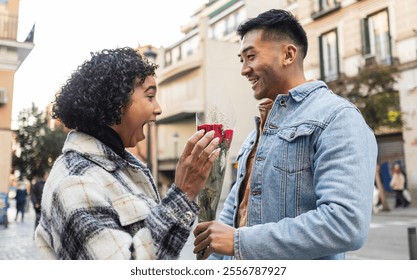 Young man is giving a red rose to a surprised young woman in the street, celebrating valentine's day with love and affection in a multiracial couple - Powered by Shutterstock