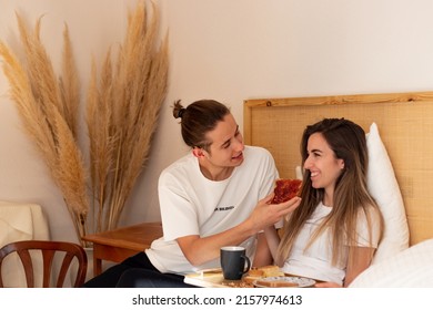 Young Man Giving Strawberry Jam Toast To His Wife In Bed While Having Breakfast In The Room. Cute Loving Couple