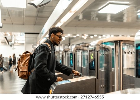 Young man with glasses and backpack tapping a contactless ticket at a modern subway turnstile, entering a bright underground station during his daily city commute