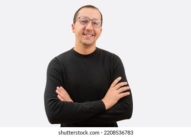 Young Man In Glasses And A Black Sweater With Crossed Arms Looking At The Camera On A White Background.
