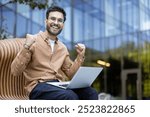 Young man with glasses sitting on bench outdoors holding laptop. Demonstrates joy and achievement. Perfect for themes of success, technology, happiness, and work-life balance.