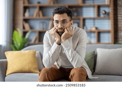 Young man with glasses sitting on sofa, deep in thought, reflecting on life choices. Dressed in casual clothes, conveying introspection and contemplation in relaxing home setting. - Powered by Shutterstock