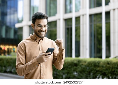 Young man in glasses smiling and celebrating success while using smartphone outdoors. Business professional receiving good news, feeling happy. Modern office building background. - Powered by Shutterstock