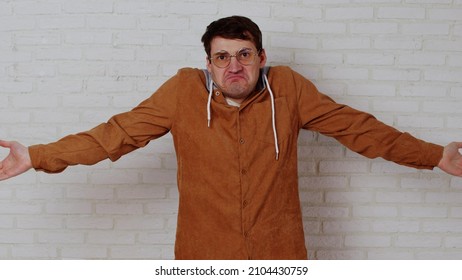 Young Man In Glasses Spreading Hands On Background Of White Brick Wall. Perplexed Male Shows Gesture Of Not Knowing, Looking At Camera