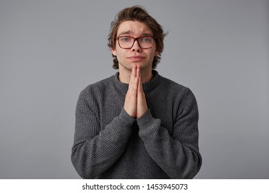 Young man with glasses wears in gray sweater, stands over gray background and looks at the camera, has sorrorful expression, keeps palms in praying gesture. - Shutterstock ID 1453945073
