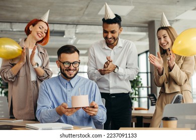 Young man is going to blow candles on cake and make a wish while celebrating birthday with colleagues. Colleagues celebrating a birthday in the office - Powered by Shutterstock
