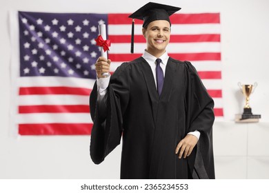 Young man in a graduation gown and cap holding a diploma in front of USA flag - Shutterstock ID 2365234553