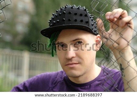 Young man with green dreadlocks in black cap, purple t-shirt posing through hole of lattice fence