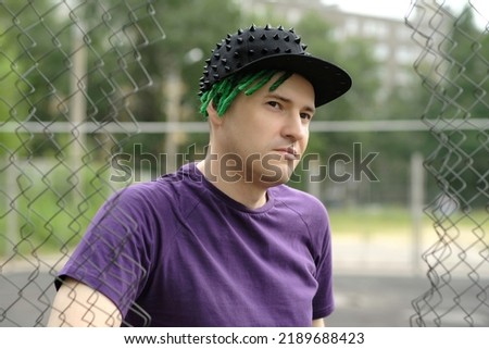 Young man with green dreadlocks in black cap, purple t-shirt posing through hole of lattice fence