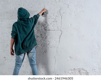 A young man in a green hoodie is spraying graffiti on a wall. Scene is rebellious and edgy, as the act of graffiti is often associated with vandalism and defiance of authority - Powered by Shutterstock