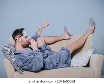 Young Man In Grey Bathrobe Sitting On The Couch Relaxing With A Glass Of Whiskey.Young Playboy In Dressing Gown Toasting With Cocktail.Drunk Man Drink Alcohol Lying In Sofa