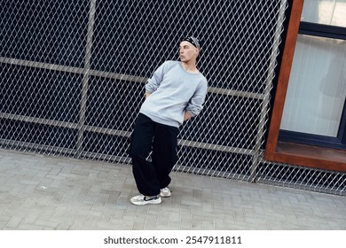 A young man in a grey sweatshirt and black pants poses in front of a chain link fence - Powered by Shutterstock