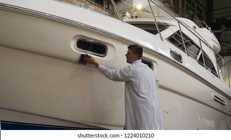 Young man (guy) specialist in white work coat polishes the yacht in the garage. Concept of: Preparation, Cleaning, Polishing, Master, Professional, After painting, Using chemistry.