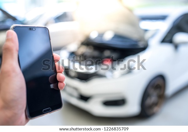 Young man hand holding and using mobile (smart\
phone) over blurred image of broken White car : concept calling\
mechanic service from repair\
shop