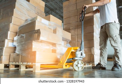Young man with hand pallet truck or pallet jack and stack of cardboard boxes on pallet in distribution warehouse.