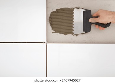 Young man hand using spatula and putting fresh glue on wall or floor concrete. Gluing white ceramic tiles. Closeup. Renovation process. Repair work of home.