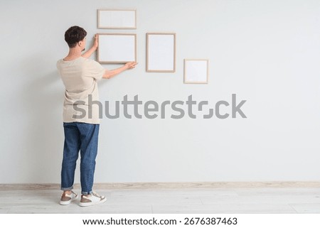 Young man hanging blank frames on light wall, back view