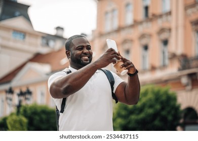 Young man happily takes a selfie with his smartphone, holding a coffee cup while exploring a european city, capturing travel moments and enjoying urban sights and sounds - Powered by Shutterstock