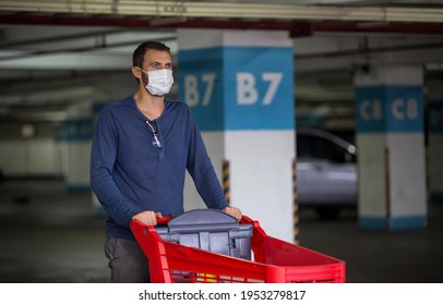 Young Man Has Medical Face Mask Against Corona Virus Covid-19 While Pushing Shopping Cart Trolley Outside Of A Grocery Store In Empty Parking Garage. Food Supplies Shortage. Panic Buying And Hoarding