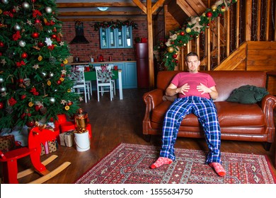 A Young Man Has A Stomach Ache Due To Overeating At A Christmas Dinner. Country Wooden House Decorated In A Christmas Style, Winter Time