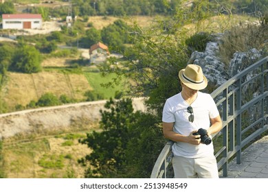 A young man in a hat adjusts the settings on a DSLR camera while standing in the mountains with a rural view in the background. The photographer takes a photo during a trip to nature on a summer day. - Powered by Shutterstock