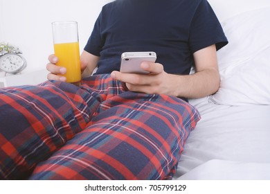Young man having breakfast and using his smartphone in bed. Indoors.