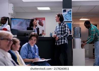 Young Man Having Casual Conversation With Hospital Receptionist At Front Desk While Nurse Is Checking Appointment With Young Girl. Asian Patient In Clinic Waiting Room With Diverse Patients.