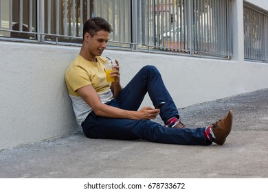 Young man having drink while using mobile phone on sidewalk by wall