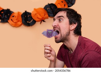 Young man is having fun, making grimaces on an isolated background with paper garland of black and orange pumpkins. The guy trying to eat a gingerbread in the form of purple bat. Halloween treat