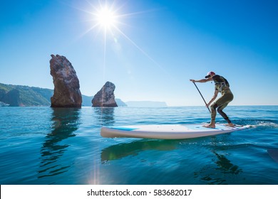 Young Man Having Fun Stand Up Paddling in the sea. SUP. Guy Training in the morning on Paddle Board near the rocks.