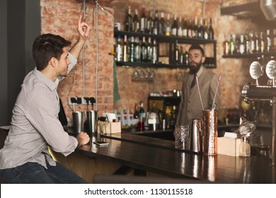 Young man having rest at friday evening, sitting at bar counter and ordering beer in bar, gesturing to bartender, copy space - Powered by Shutterstock