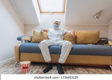 Young Man In Hazmat Suit And Protective Mask Holding Bottle Of Beer While Sitting On Sofa Near Popcorn Bucket On Floor