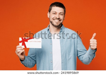 Young man he wears blue shirt white t-shirt casual clothes hold gift certificate coupon voucher card for store show thumb up isolated on plain red orange background studio portrait. Lifestyle concept