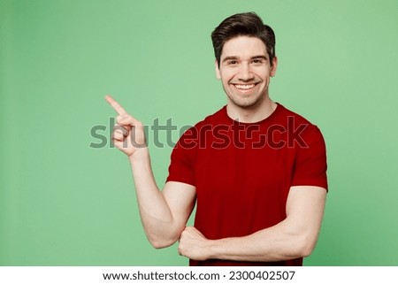 Young man he wears red t-shirt casual clothes point index finger aside indicate on workspace area copy space mock up isolated on plain pastel light green background studio portrait. Lifestyle concept