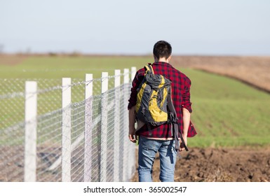 Young man with head down and backpack on the shoulder walking beside wire fence. Rear view of man