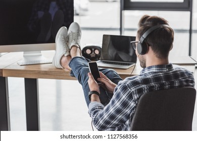 Young Man In Headphones Using Smartphone While Sitting With Legs On Table With Laptop And Desktop Computer