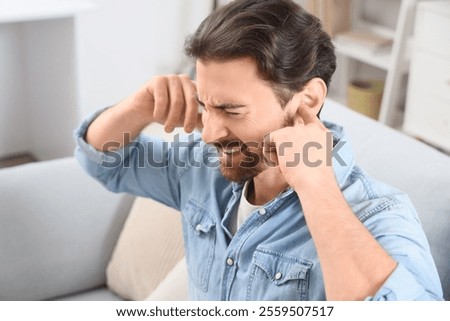 Young man with hearing problem covering his ears on sofa at home, closeup