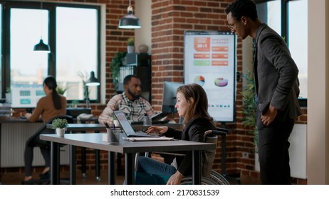 Young Man Helping Businesswoman In Wheelchair To Work At Desk, Giving Support In Office. Female Worker With Disability Planning Leadership Analysis With Financial Data Charts And Diagram.