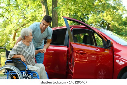 Young Man Helping Disabled Senior Woman In Wheelchair To Get Into Car Outdoors